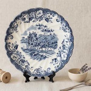 May include: A blue and white decorative plate with a scalloped edge. The plate features a pastoral scene with a horse-drawn carriage and floral border. The plate is displayed on a black stand.