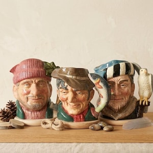 May include: Three ceramic mugs shaped like men's heads, each with a unique hat and facial expression. One wears a pink knit cap, another a brown hat, and the third a striped hat. Each mug has a different accessory.