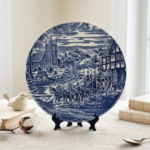 May include: A blue and white decorative plate featuring a pastoral scene with a horse-drawn carriage, church, and village. The plate is displayed on a black stand, with a bird figurine and books in the background.