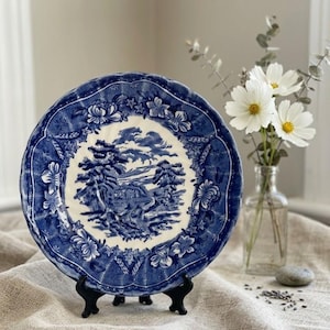 Può includere: Un piatto decorativo blu e bianco con una scena di paesaggio al centro. Il piatto ha un bordo smerlato con motivi floreali. È esposto su un supporto nero. Fiori bianchi in un vaso di vetro sullo sfondo.
