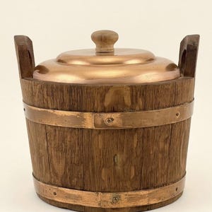 May include: A wooden bucket with a copper lid and bands. The bucket has two wooden handles and a small wooden knob on top of the lid. The wood has a natural grain, and the copper has a shiny, reflective surface.