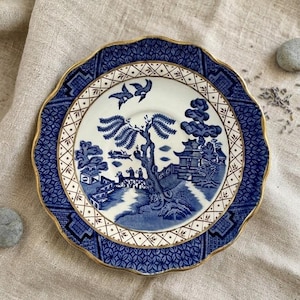 Puede incluir: Un plato de porcelana azul y blanco con borde dorado. El plato presenta una escena detallada de paisaje con un puente, árboles y edificios. El borde exterior tiene un diseño estampado. El plato tiene unos 20 cm de diámetro.
