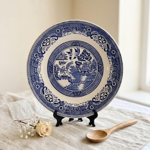 May include: A blue and white ceramic plate with a detailed Willow pattern design. The plate is displayed on a black stand, with a wooden spoon and a white rose nearby. The plate's rim features a repeating geometric pattern.