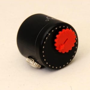 Può includere: Un oggetto cilindrico nero con un componente rosso a forma di ingranaggio. Ha un piccolo pomello argentato e una serie di punti bianchi. L'oggetto è su uno sfondo bianco. Il componente rosso ha una piccola freccia.