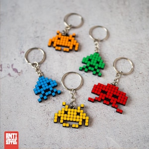 Puede incluir: Cinco llaveros alienígenas pixelados en naranja, verde, azul, amarillo y rojo. Cada llavero presenta un anillo plateado y un pequeño diseño alienígena colorido. Los llaveros están dispuestos sobre una superficie gris claro.