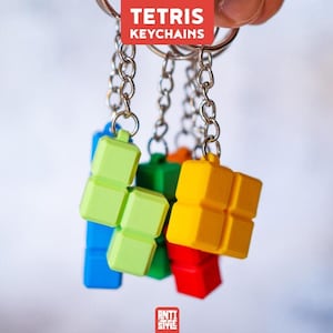 May include: A collection of Tetris keychains featuring colorful plastic geometric blocks in yellow, lime green, red, blue, and orange. Each block is attached to a silver metal chain and ring. The text TETRIS KEYCHAINS is visible at the top.