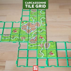 May include: A green plastic grid frame for board game tiles, featuring a modular design with empty square slots and several square tiles placed within, depicting illustrated landscapes with roads, fields, and buildings. Text reads CARCASSONNE TILE GRID.