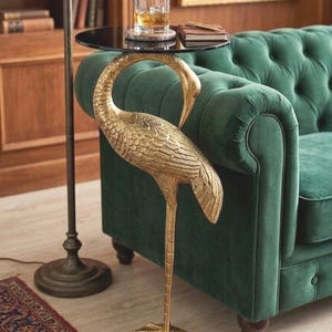 Könnte beinhalten: Ein goldfarbener Beistelltisch mit Kranich-Design und schwarzer Glasplatte. Der Tisch hat eine detaillierte Kranich-Skulptur als Basis. Er steht neben einem grünen Samtsofa und einer Stehlampe, was eine luxuriöse Wohnatmosphäre schafft. Die Höhe beträgt ca. 70 cm.