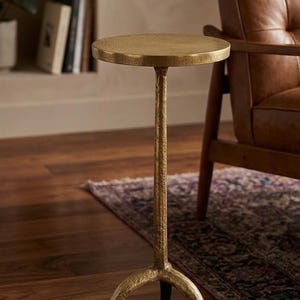 Puede incluir: Una mesa auxiliar dorada con una tapa redonda y un tallo delgado y texturizado. La base tiene tres patas curvas. La mesa es de metal y es adecuada para una sala de estar o un dormitorio.