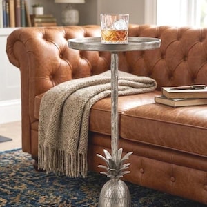 May include: A silver-colored side table with a pineapple base and a round top. A glass of amber liquid with ice sits on the table. A brown leather sofa and a patterned rug are in the background.
