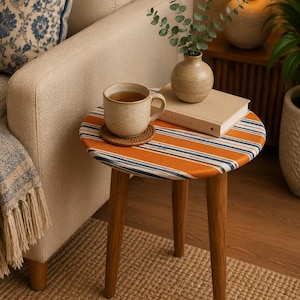 May include: A round side table with three tapered wooden legs and a top upholstered in orange, blue, and white striped fabric. On the table sits a speckled ceramic mug on a woven coaster, a beige book titled The Kinfolk, and a small ceramic vase.