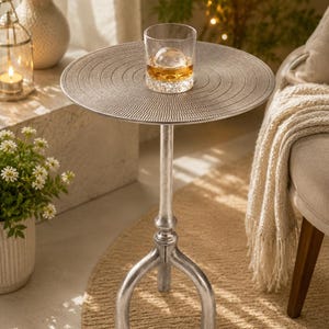May include: A silver-colored side table with a round top and three curved legs. The table has a textured surface and supports a glass of amber liquid with a large ice sphere. Decorative home decor.