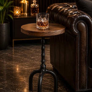 Puede incluir: Una mesa auxiliar redonda de madera con una superficie de veta marrón oscuro y una base de metal negro con tres patas curvas. Un vaso de cristal transparente con líquido ámbar y cubitos de hielo descansa en el centro de la mesa.