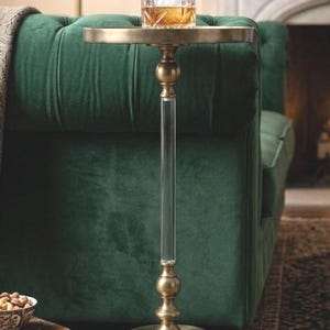 Puede incluir: Una mesa auxiliar dorada con un tallo de acrílico transparente y una tapa redonda. Un vaso de líquido ámbar con hielo descansa sobre la mesa, junto a un sofá de terciopelo verde.
