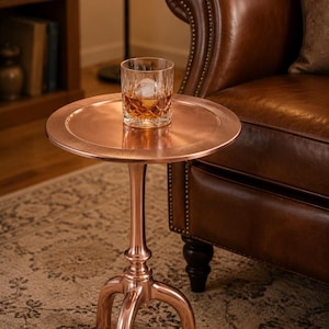 May include: A round copper-toned metal side table with a smooth top surface and a textured edge, supported by a central pedestal base that splits into three curved legs. A clear glass tumbler containing an amber liquid and a round ice sphere sits on top.
