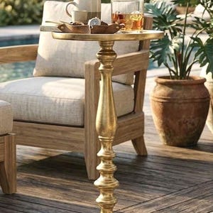 May include: A gold-colored, ornate side table with a round top. The table is outdoors, with a wooden chair and a potted plant in the background. The table holds a cup, a small dish, and a glass of amber liquid.