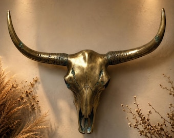 May include: A bronze-colored bull skull wall decor with curved horns. The skull has a detailed texture and is mounted on a neutral-toned wall. The horns are long and curved, adding a rustic touch to the piece.