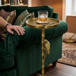 May include: A brass-coloured side table with a parrot design, holding a glass of amber liquid with a large ice sphere. The table is next to a dark green velvet sofa. The table's height is approximately 60 cm.