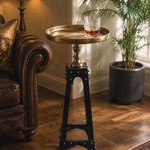 May include: A decorative side table with a round, antique brass tray top. The table has a black, industrial-style base and a gold-colored pedestal. A glass of amber liquid sits on the tray. The table is in a living room setting.