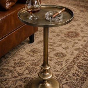 May include: A round, antique brass-finished metal side table with a slender pedestal base. On the tabletop sits a clear glass snifter filled with amber liquid and a clear glass ashtray containing a single brown cigar.