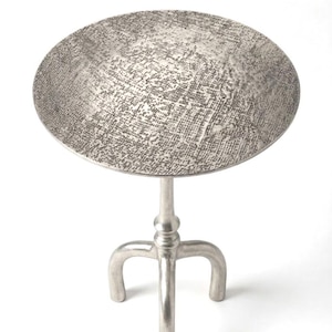 Può includere: Tavolino laterale argentato con piano rotondo e tre gambe curve. Il piano del tavolo ha una superficie strutturata e decorata. Il tavolo è in metallo e si trova su uno sfondo bianco.