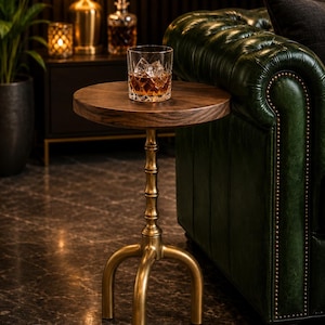 May include: A round wooden side table with a dark brown grain top and a metallic gold-toned base featuring a central column and three curved legs. A clear glass tumbler containing an amber liquid and ice cubes sits on the table surface.