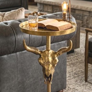 Könnte beinhalten: Ein goldfarbener Beistelltisch mit einem Bullenschädel-Design. Der Tisch hat eine runde Tablettplatte, eine zentrale Stütze und einen Holzsockel. Ein Glas Whisky, ein Buch und eine kleine Lampe stehen auf dem Tisch.