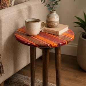 May include: A round side table with three dark brown wooden legs and a tabletop featuring a vibrant orange, red, and yellow patterned design. The table surface holds a speckled ceramic mug, a small ceramic vase with green leaves, and a book.