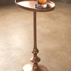 May include: A copper-colored side table with an oval top and base. The table has a decorative, ornate stem. A glass of amber liquid with a large ice sphere sits on the table, resting on a coaster.