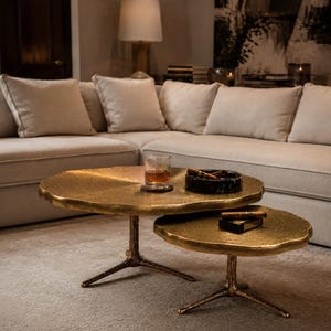 May include: A set of two round nesting coffee tables with textured gold-toned surfaces and tripod bases shaped like tree branches. The tables feature irregular, organic edges and a metallic finish, providing a functional surface for small items.