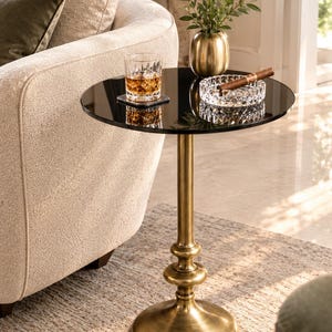 Puede incluir: Una mesa auxiliar redonda con una superficie de cristal negro reflectante y una base de pedestal de latón cepillado. La mesa presenta una columna central con detalles redondeados decorativos y una base circular ancha que sostiene un vaso.