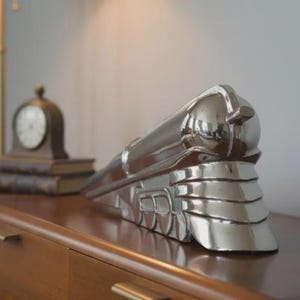 Puede incluir: Una escultura de tren plateada pulida, un objeto decorativo, se encuentra sobre una superficie de madera. El tren está detallado con un diseño aerodinámico. Un pequeño reloj y libros están en el fondo.