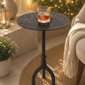 Könnte beinhalten: Ein schwarzer Beistelltisch aus Metall mit strukturierter Tischplatte. Ein Glas bernsteinfarbener Flüssigkeit mit Eis und einer Orangenschale steht auf dem Tisch. Der Tisch hat ein dreibeiniges Gestell und steht auf einem gewebten Teppich.