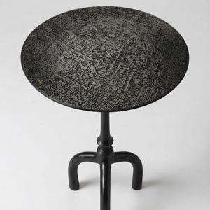 Può includere: Un tavolino nero con piano rotondo e tre gambe curve. Il piano del tavolo ha una superficie strutturata e decorata. Il tavolo è alto circa 61 cm e il piano misura 46 cm di diametro.