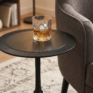 May include: A glass of amber liquid with a large ice sphere sits on a black side table. The glass has a patterned design. The table has a textured rim and a single, black pedestal base. A grey armchair is in the background.