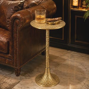 Puede incluir: Una mesa auxiliar redonda de metal dorado con una superficie texturizada y martillada en el tablero, el pedestal central y la base circular. La mesa sostiene un vaso transparente con líquido ámbar y un pequeño cenicero de metal para puros.