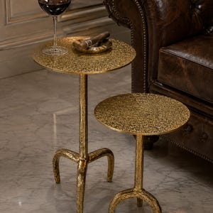 Puede incluir: Dos mesas auxiliares redondas con acabado dorado texturizado y tres patas curvas. La mesa más alta sostiene una copa de vino tinto y un cigarro en un cenicero. Las mesas presentan una superficie de metal martillado y un diseño de base a juego.