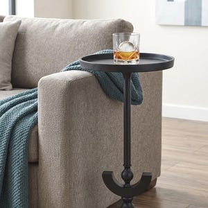 May include: A black side table with a round top and a decorative base. A glass of amber liquid with a large ice sphere sits on the table. A blue blanket is draped over the arm of a beige sofa.