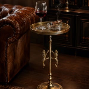 Puede incluir: Una mesa auxiliar redonda de latón con una base de pedestal que presenta dos figuras de leones esculpidas trepando por la columna central. La superficie sostiene una copa de vino con vino tinto y un cenicero de cristal con un cigarro encendido.