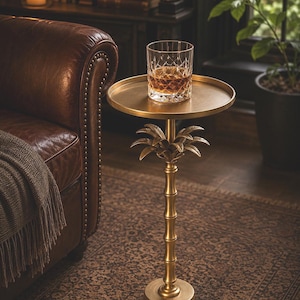 Puede incluir: Una mesa auxiliar dorada con base de diseño de palmera, sosteniendo un vaso de cristal lleno de líquido ámbar. La mesa tiene una parte superior redonda y un tallo estilo bambú. El entorno incluye un sillón de cuero y una alfombra estampada.