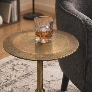 Pode incluir: Um copo de vidro transparente com padrão de diamante contendo um líquido âmbar e um cubo de gelo esférico, sobre uma mesa lateral redonda de metal com acabamento em latão antigo, base central e três pernas curvas.