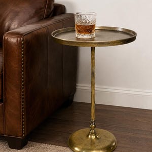 Puede incluir: Una mesa auxiliar redonda de metal con acabado en latón antiguo, que presenta un pedestal central texturizado y una base circular ancha. Un vaso de cristal tallado transparente con un líquido de color ámbar descansa sobre la superficie plana.