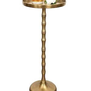 May include: A gold-colored side table with a round tray top. The table has a bamboo-style stem and a circular base. A glass of amber liquid with ice and a white flower rest on the tray.