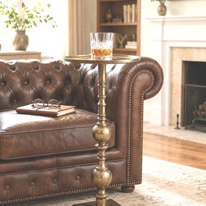 May include: A brass side table with a round top and an ornate stem. A glass of amber liquid sits on the table. The table is next to a brown leather sofa. The table's height is approximately 76 cm.