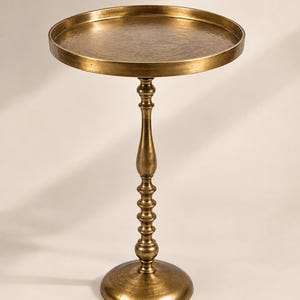 Op de afbeelding: Een ronde bijzettafel van messing met een gehamerde textuur op het blad. De tafel heeft een hoge, slanke centrale kolom met decoratieve gedraaide details en een brede, ronde voet voor stabiliteit, afgewerkt in een warme metaalkleur.
