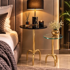 May include: Two round side tables with textured gold-tone metal tripod bases. The taller table features a black circular top holding a black marble-based lamp with a black shade, a reed diffuser, and a candle. The shorter table has a clear glass top.
