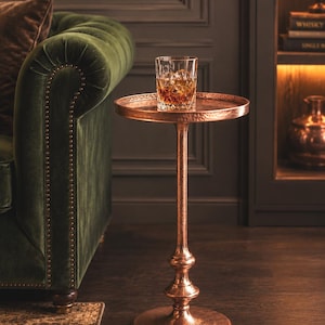 May include: A tall, round side table with a hammered copper finish, featuring a slender pedestal base and a circular top. A clear glass tumbler filled with an amber-coloured liquid and ice cubes sits centred on the table surface.