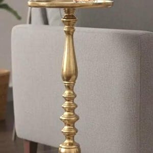 May include: A gold-colored side table with a round top and ornate, detailed pedestal. A glass of amber liquid sits on the table. The table is placed next to a gray sofa, suggesting a living room setting.