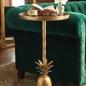 May include: A gold-colored side table with a pineapple base and a round top. The table holds an open book, a small decorative box, glasses, and a vase with a white flower. The table is placed next to a green velvet sofa.