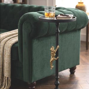 May include: Black side table with a round top and a decorative gold-colored monkey climbing the pole. The table holds a glass of amber liquid, a notebook, and a pen. The table is next to a green velvet sofa.